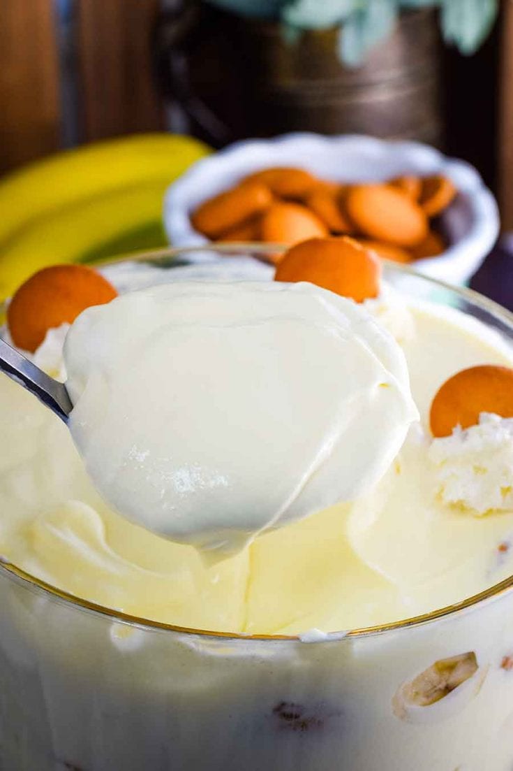 A large glass serving bowl filled with this easy banana pudding recipe topped with whipped cream and a vanilla wafers and a serving spoon scooping out a portion