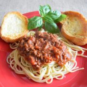 a red plate with a serving of this spaghetti with meat sauce aka bolognese sauce, fresh basil, and crispy toasted bread