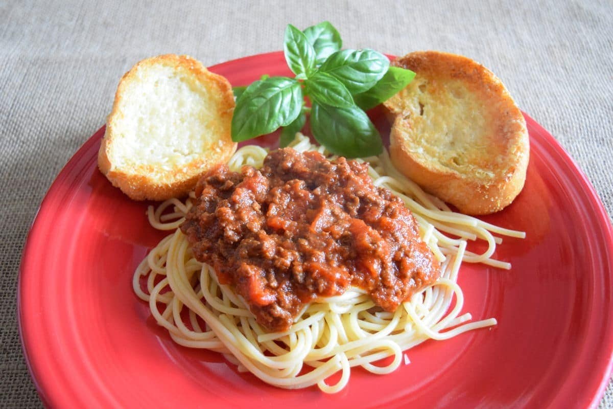 a red plate with a serving of this spaghetti with meat sauce aka bolognese sauce, fresh basil, and crispy toasted bread