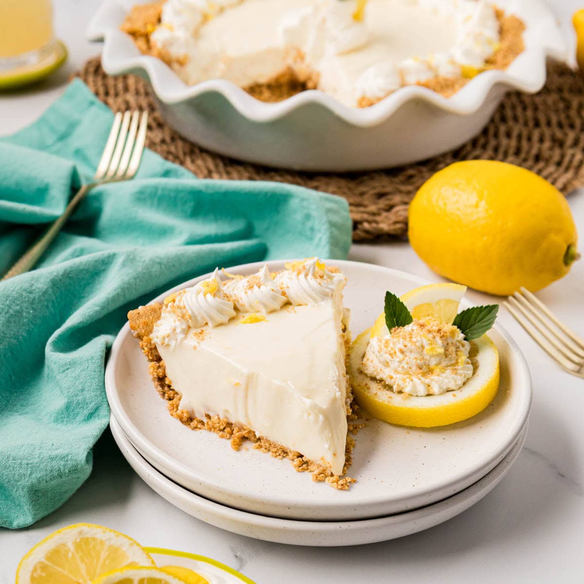 A slice of lemon icebox pie on a white plate with a lemon slice, whipped cream and mint leaves with the cut pie in the background.