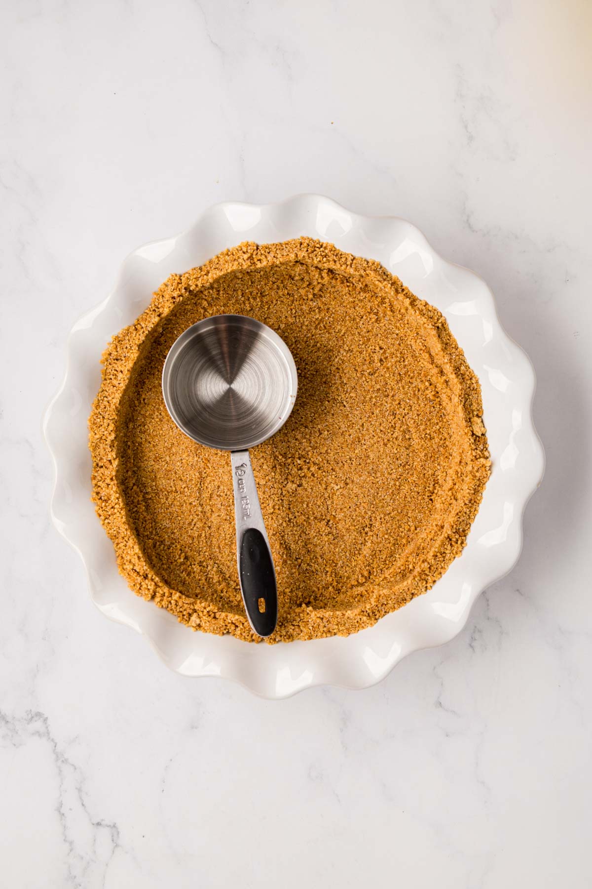 A graham cracker crust being pressed into a pie plate with a measuring cup.