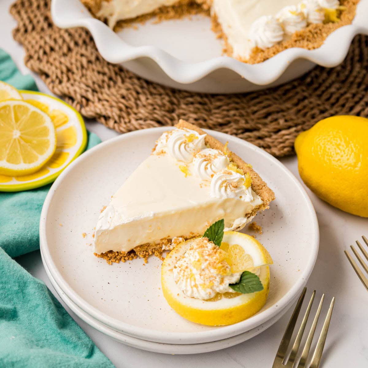 A slice of lemon icebox pie on a white plate with a lemon slice, whipped cream and mint leaves with the cut pie in the background.