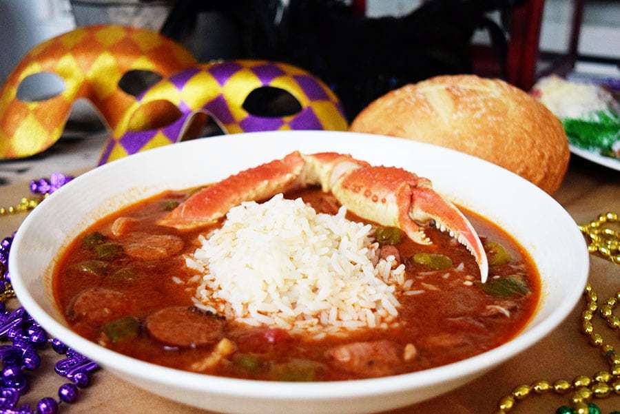 a serving of this cajun seafood and sausage gumbo in a white bowl with white rice and a cooked crab leg, as well as crusty bread and a mardi gras mask in the background