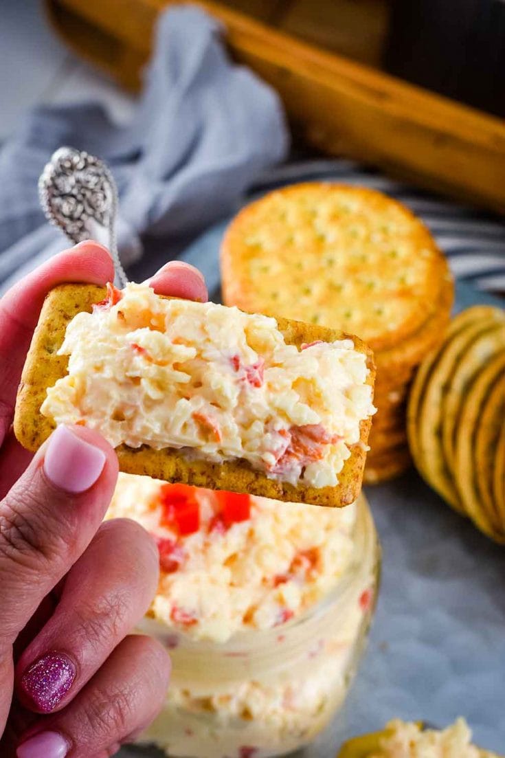 a cracker topped with small glass jar filled with this Southern Pimento Cheese with a serving spoon and some crackers in the background