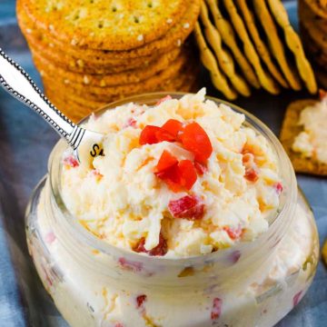 a small glass jar filled with this Southern Pimento Cheese with a serving spoon and some crackers in the background