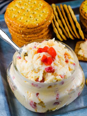 a small glass jar filled with this Southern Pimento Cheese with a serving spoon and some crackers in the background