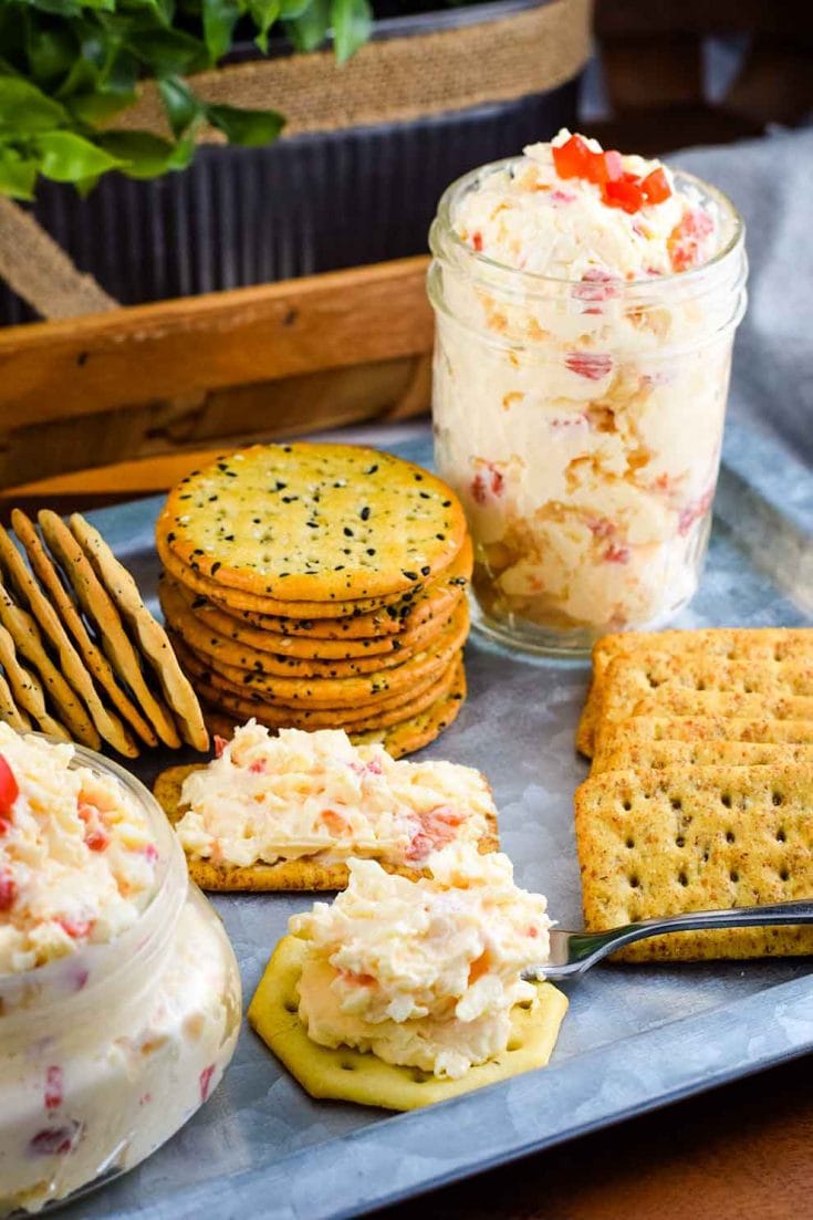 crackers topped with small glass jar filled with this Southern Pimento Cheese with a serving spoon, a jar of pimento cheese, some crackers in the background