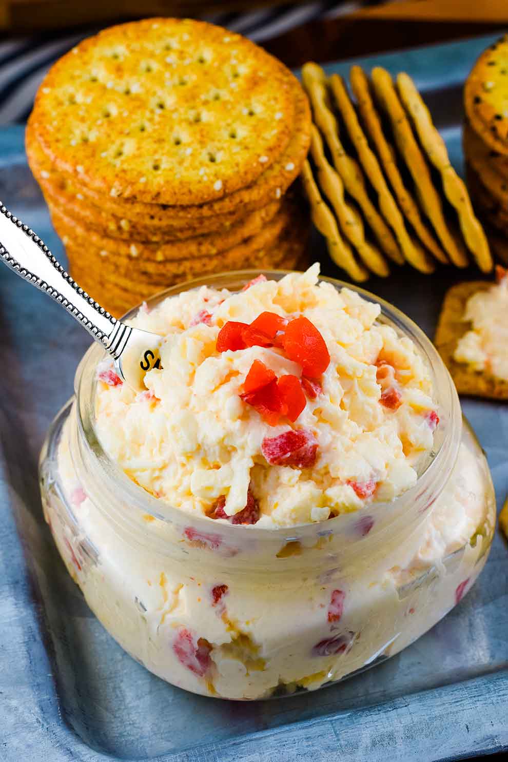 a small glass jar filled with this Southern Pimento Cheese with a serving spoon and some crackers in the background