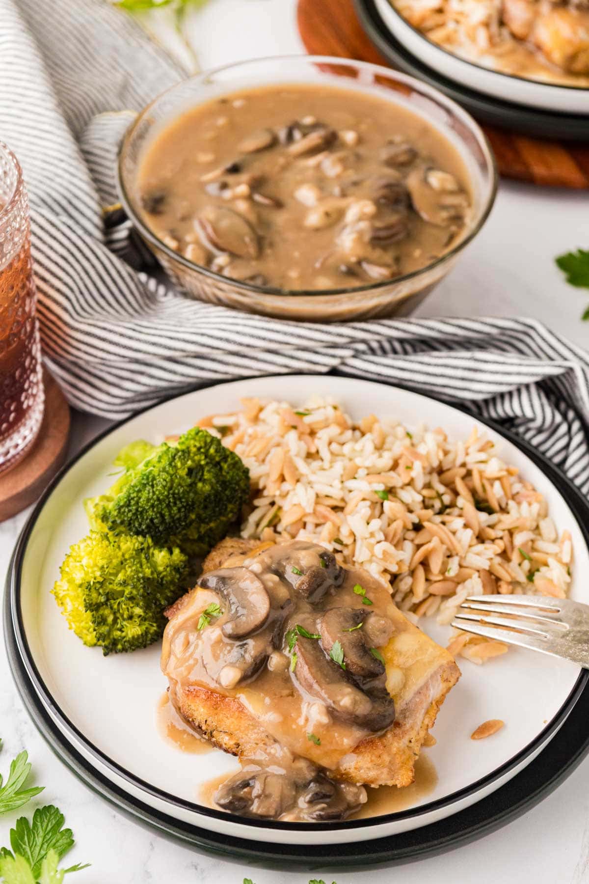 Pork chops topped with fontina cheese and a marsala mushroom sauce on a white serving plate with rice, peanut and broccoli with a bowl of the mushroom sauce in the background.