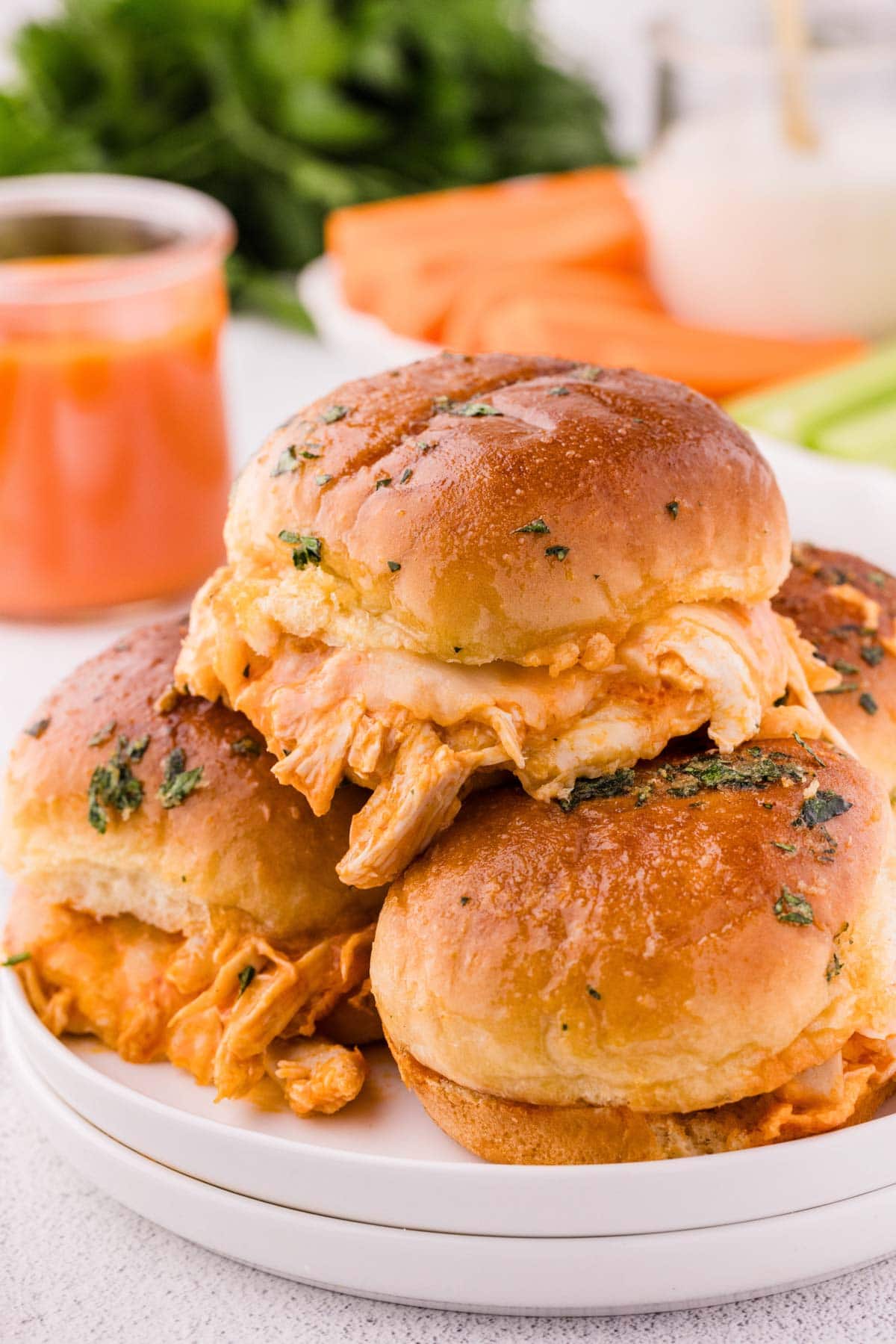 A white plate stacked with buffalo chicken sliders with carrot, celery, ranch and buffalo sauce in the background.