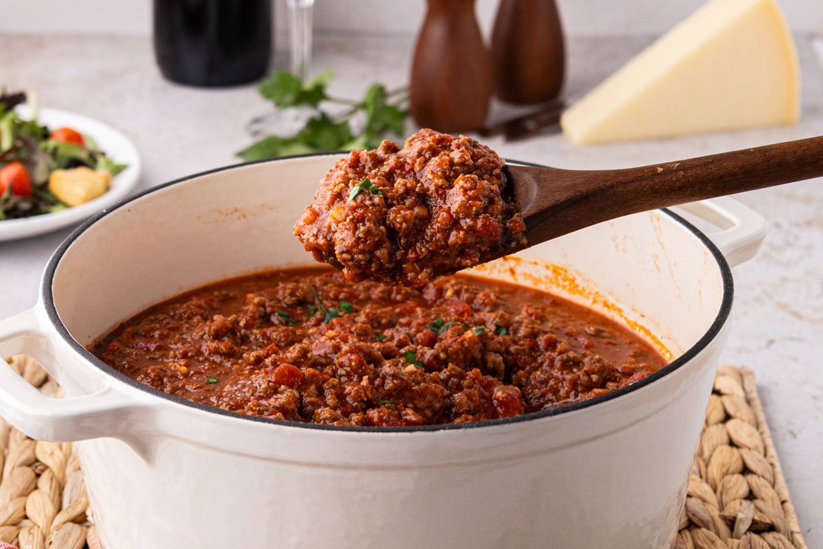 Homemade meat sauce in a white dutch oven with a wooden spoon removing a scoop.
