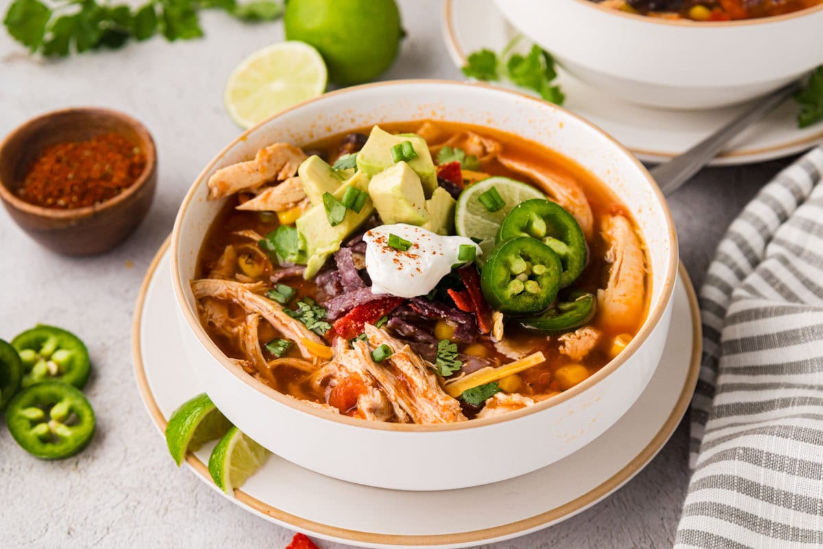 A bowl of homemade chicken tortilla soup topped with a dollop of sour cream, diced avocado, sliced jalapeños, and tortilla strips.