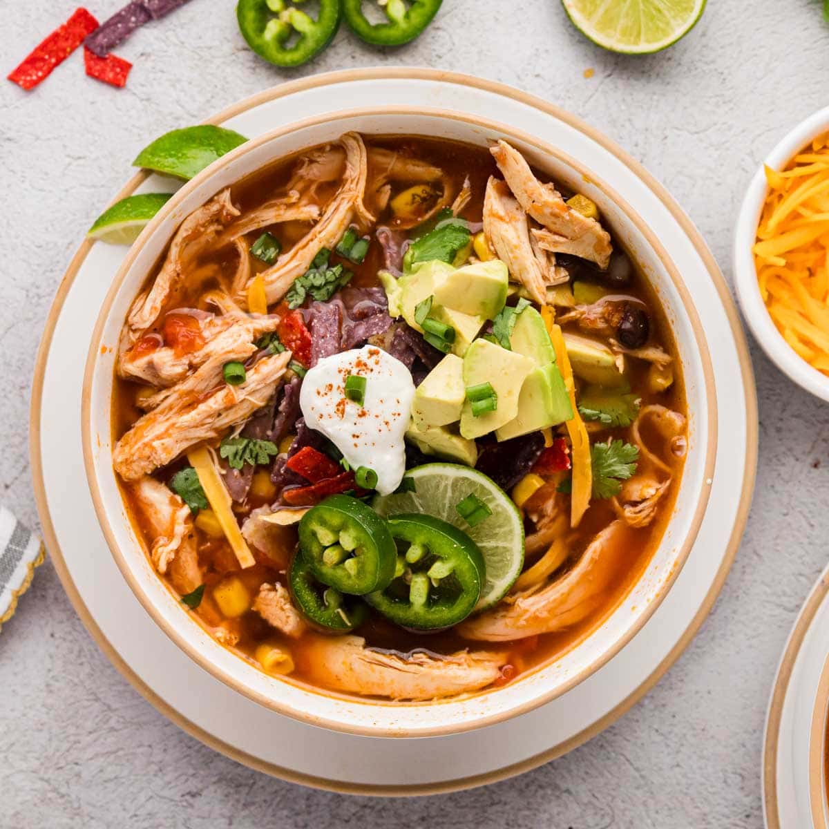 An overhead image of a bowl of homemade chicken tortilla soup topped with a dollop of sour cream, diced avocado, sliced jalapeños, and tortilla strips.