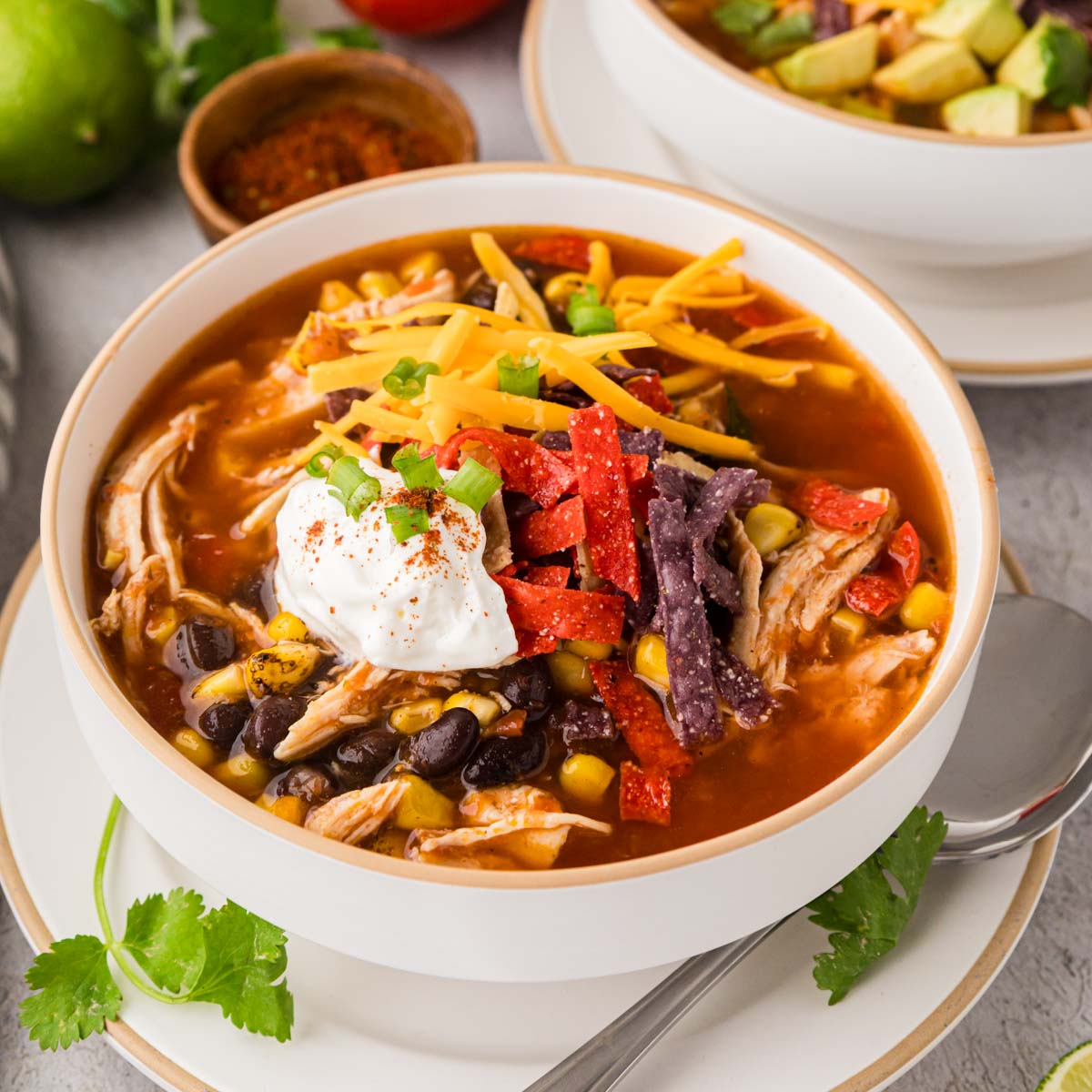 A white soup bowl filled with homemade chicken tortilla soup topped with shredded cheese, a dollop of sour cream and tortilla strips.