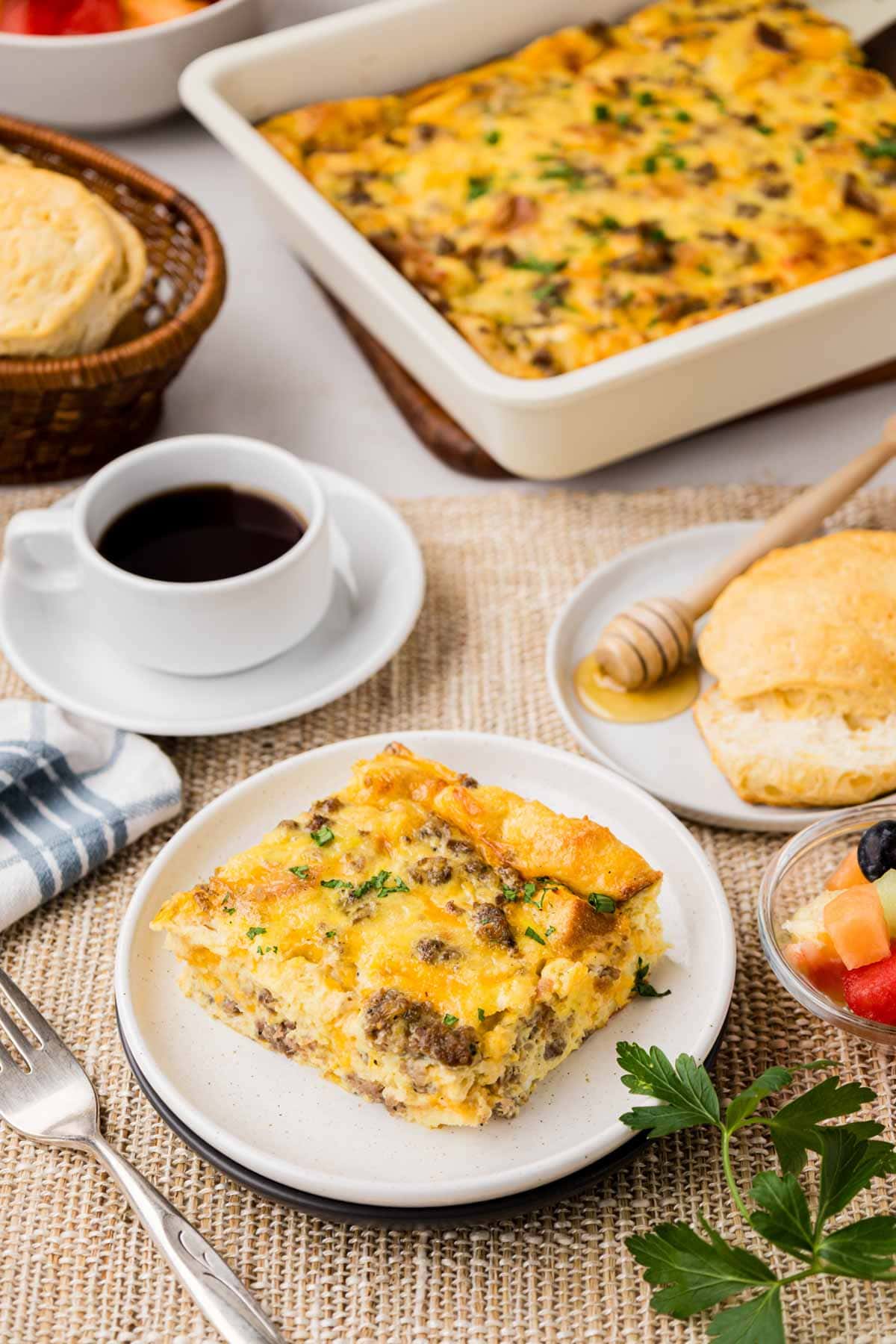 A slice of overnight breakfast casserole on a white plate set on a table with coffee, fruit, a biscuit and the casserole in the background.
