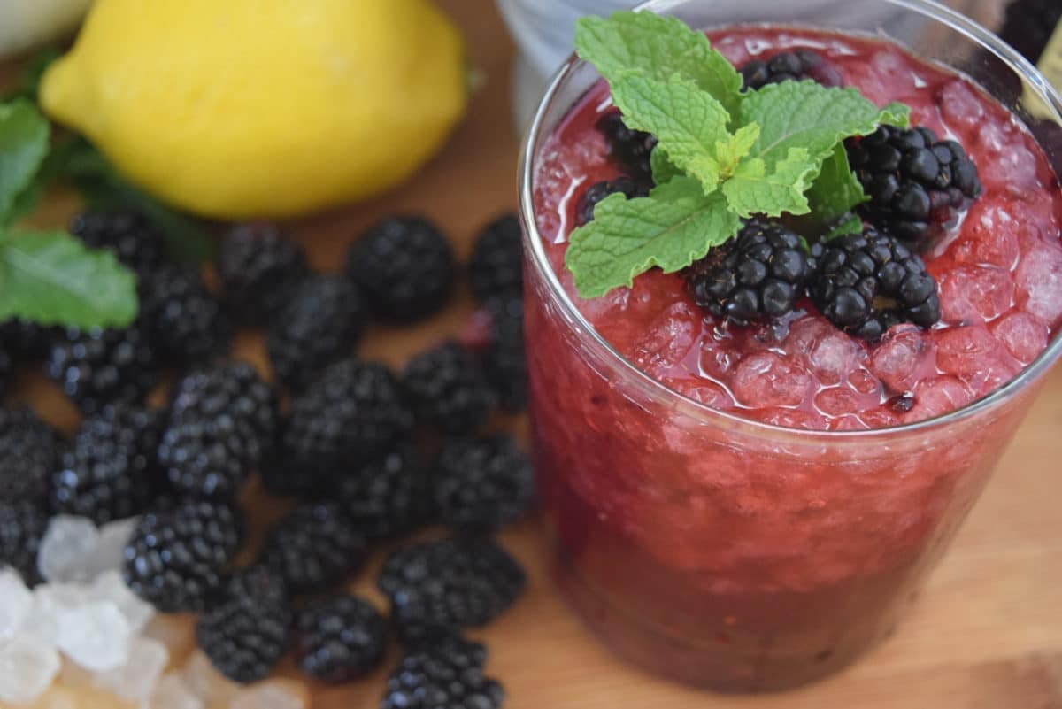 Blackberry Bramble Wish Cocktail in a glass with crushed ice on a wooden tray with blackberries, lemon and mint  in the background.