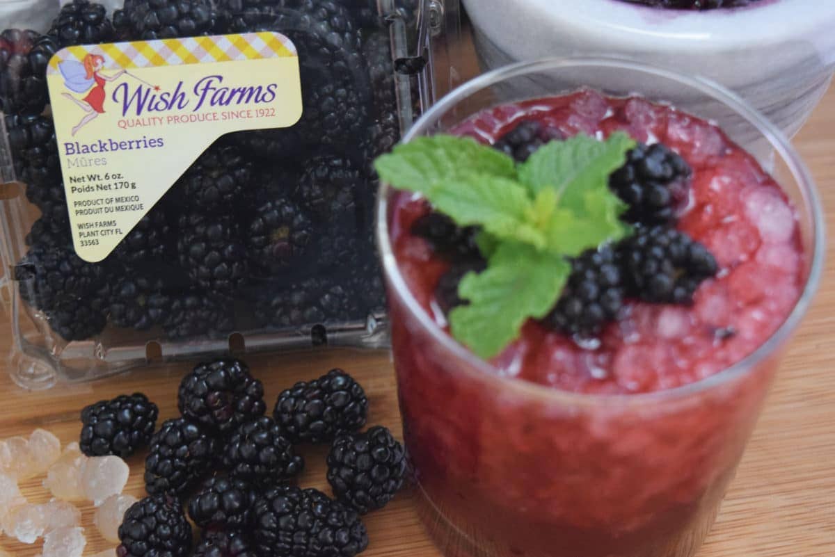 Blackberry Bramble Wish Cocktail in a glass with crushed ice on a wooden tray with wish farms blackberry container in the background.