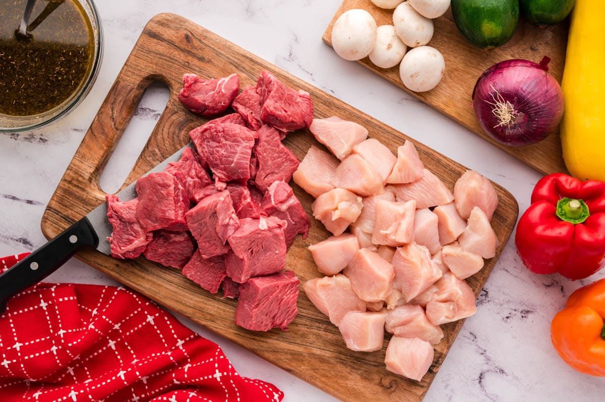 A knife on a cutting board with cubed cut pieces of steak and chicken.