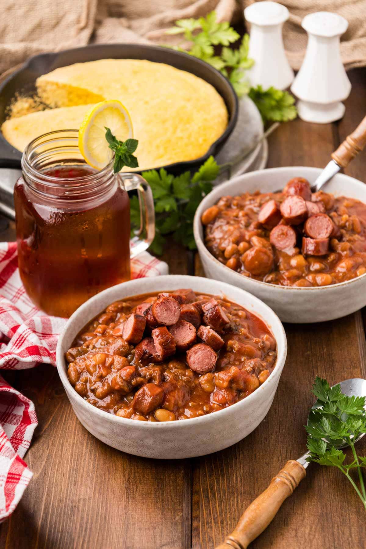 Two white serving bowls full of franks and beans with a pan of skillet cornbread in the background.