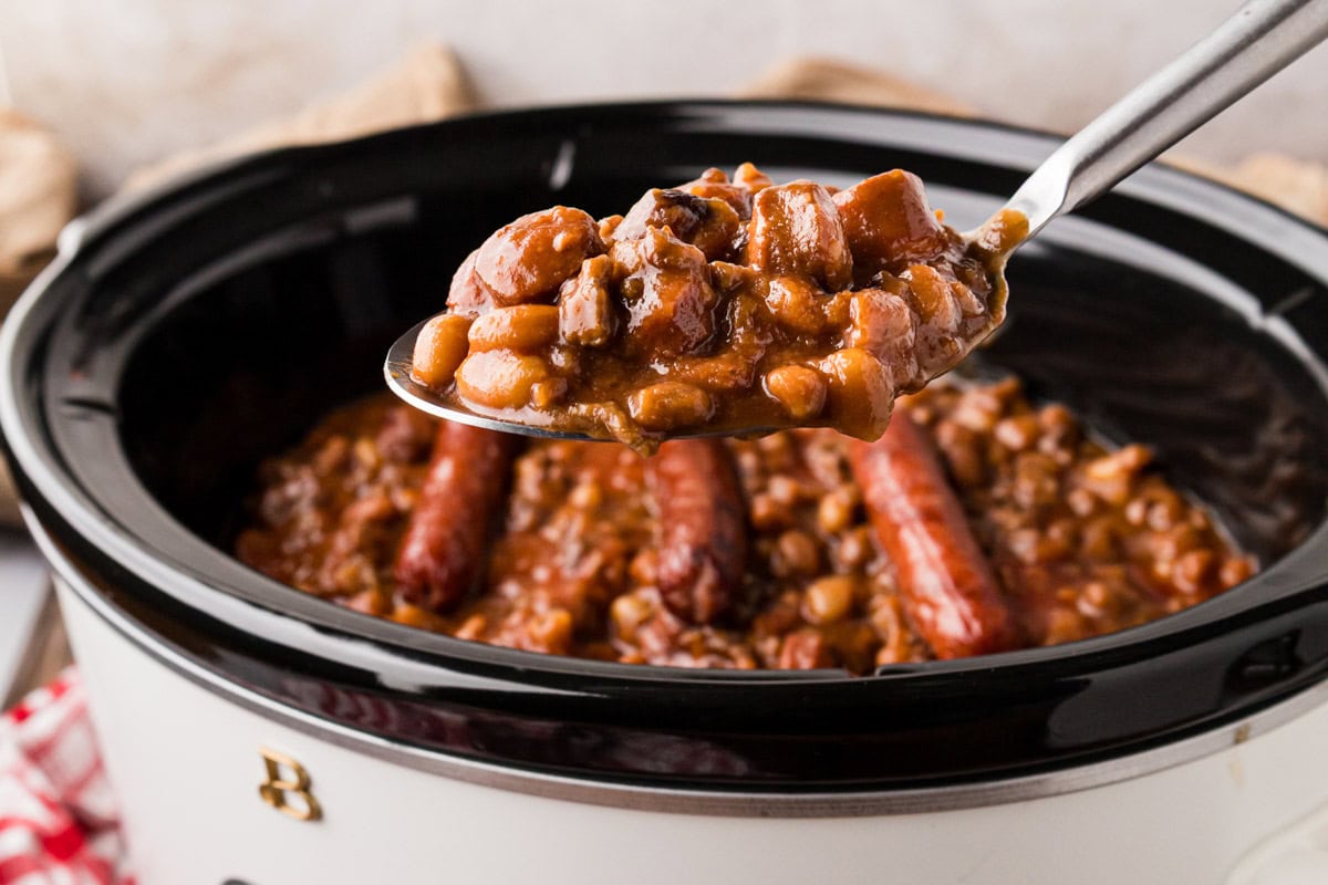 A spoon removing a scoop of beanie weenies from a slow cooker.