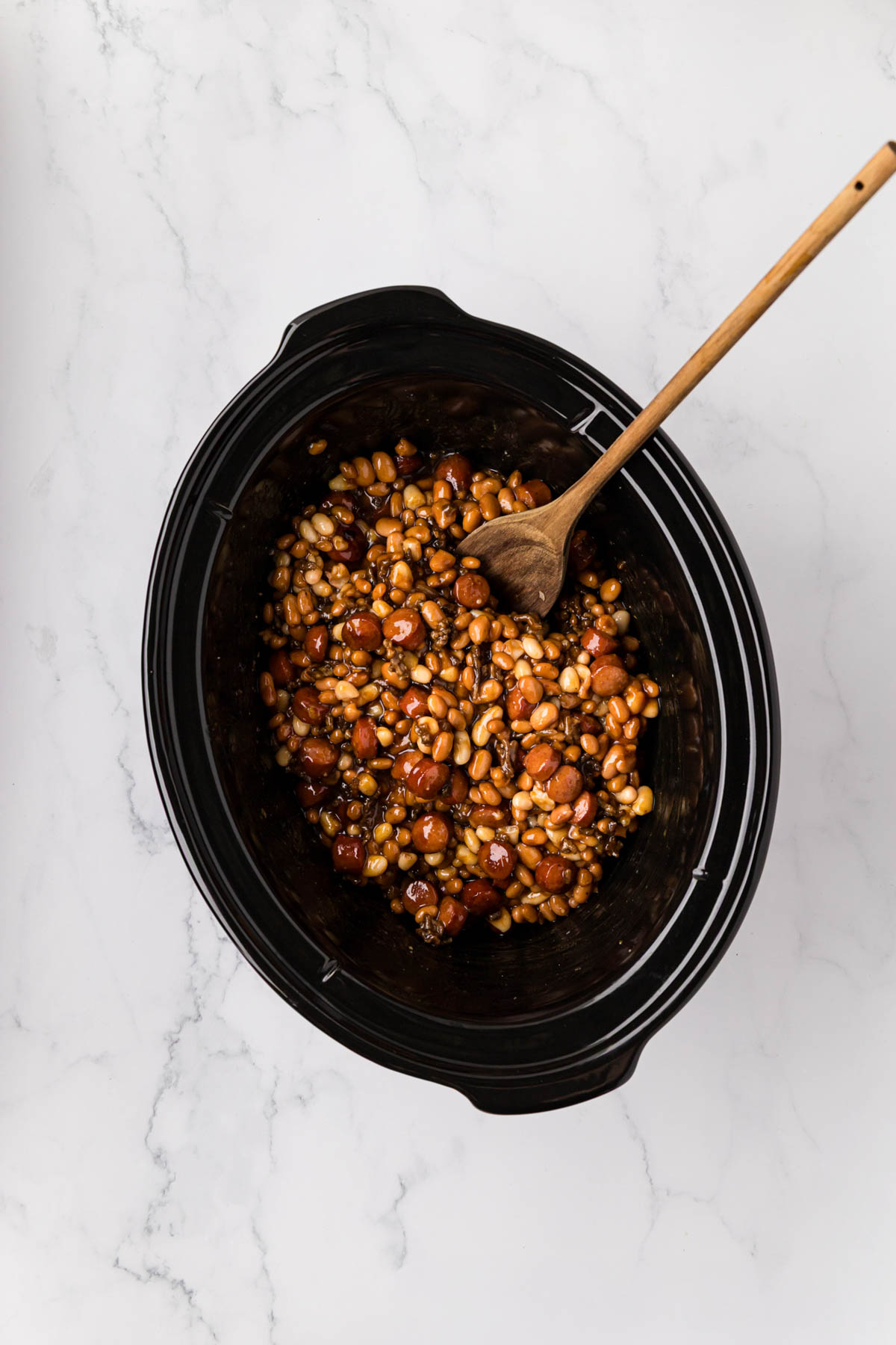 A wooden spoon stirring Ingredients to make franks and beans in a slow cooker.
