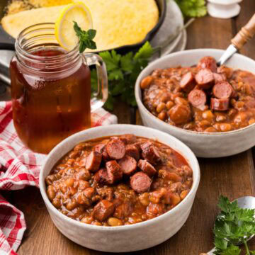 Two white serving bowls full of franks and beans with a pan of skillet cornbread in the background.