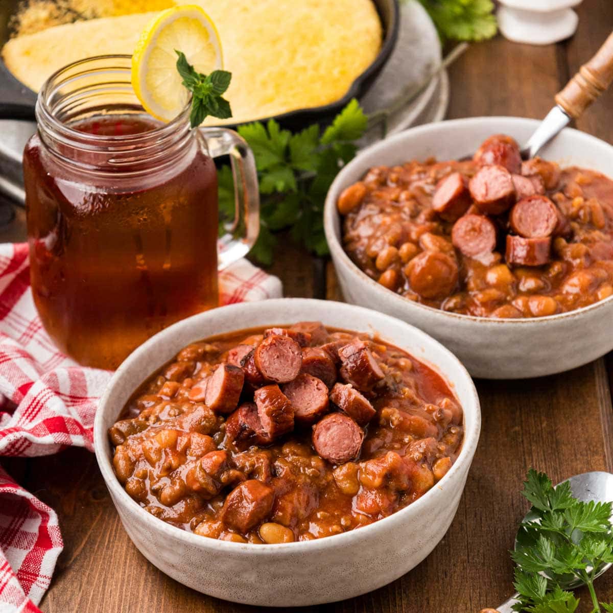 Two white serving bowls full of franks and beans with a pan of skillet cornbread in the background.