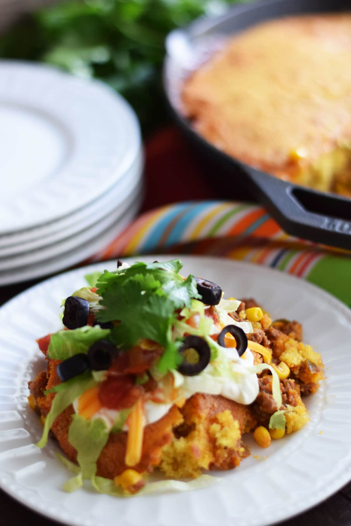 a closeup of a serving of this jalapeno cornbread taco pie on a white plate topped with black olives, salsa, shredded cheese, and shredded lettuce
