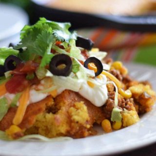 a closeup of a serving of this jalapeno cornbread taco pie on a white plate topped with black olives, salsa, shredded cheese, and shredded lettuce