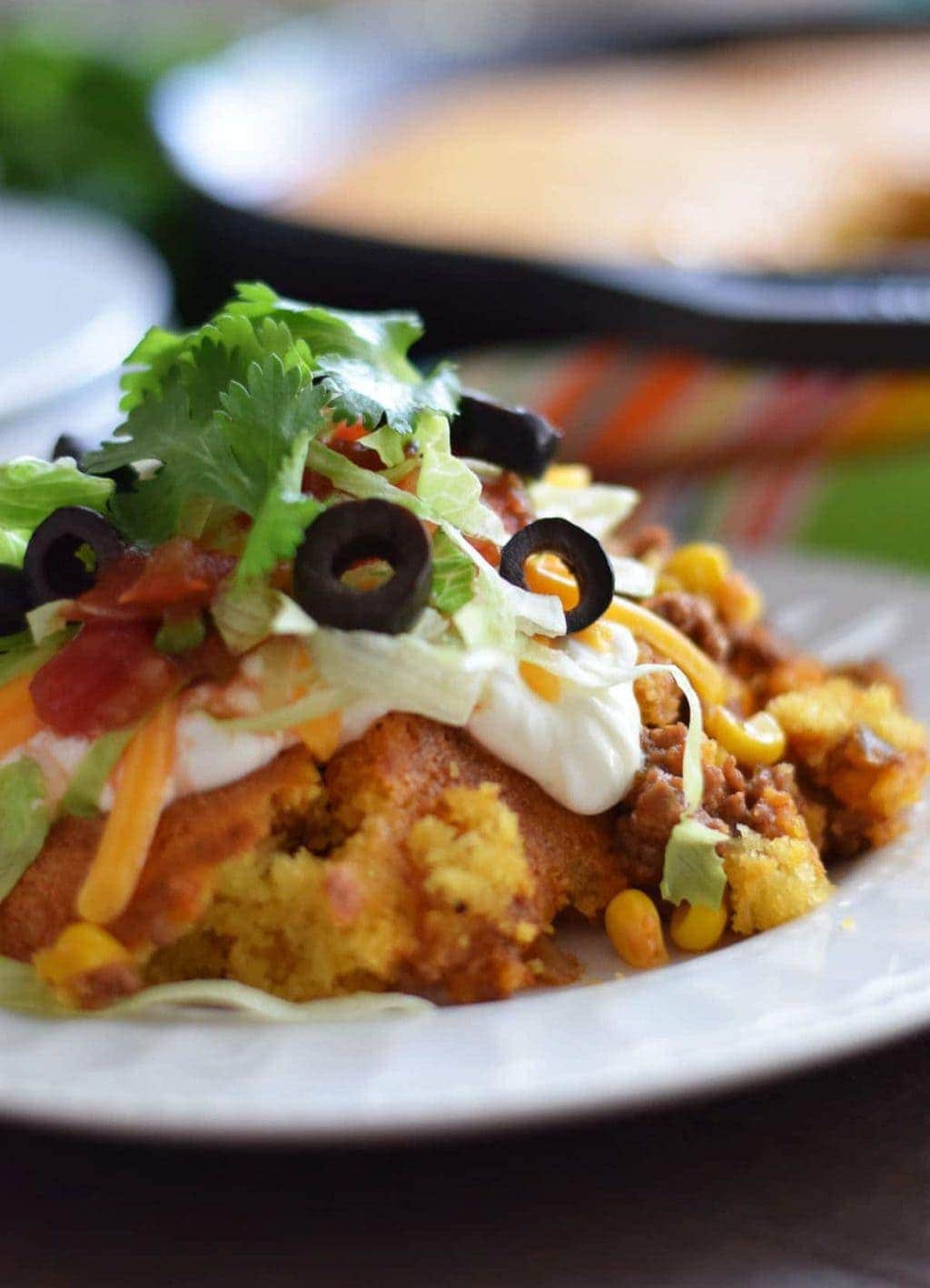 a closeup of a serving of this jalapeno cornbread taco pie on a white plate topped with black olives, salsa, shredded cheese, and shredded lettuce