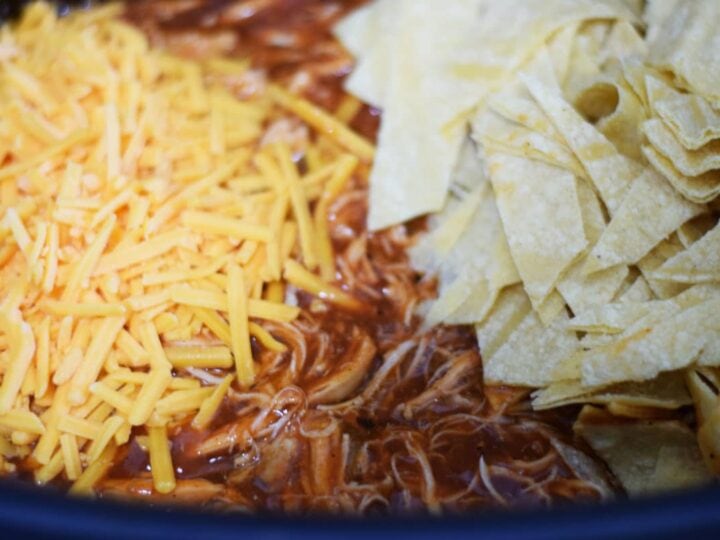 a closeup of the shredded chicken, enchilada sauce, shredded cheese, and tortilla strips inside of a slow cooker for this crock pot chicken enchilada casserole recipe