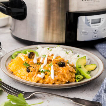 A plate with enchilada casserole made in the slow cooker, rice with black olives and avocado and jalapeño slices.