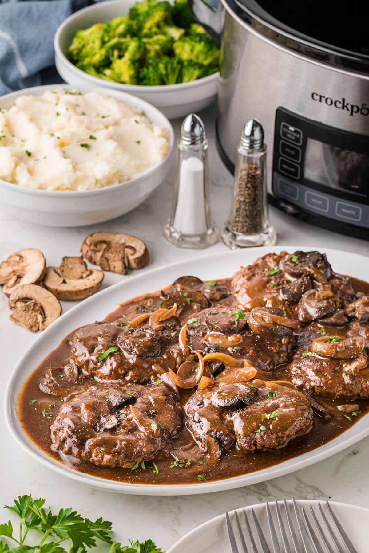 A white platter filled with Salisbury steak with a crockpot, mashed potatoes and salt and pepper in the background.