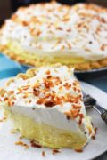 A closeup of a slice of this homemade coconut cream pie topped with shredded toasted coconut and a full coconut cream pie in the background