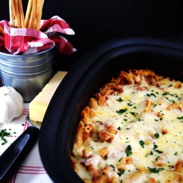 an overhead view of this crock pot chicken parmesan penne pasta with a wedge of parmesan, a cheese grater, and thin bread sticks in the background