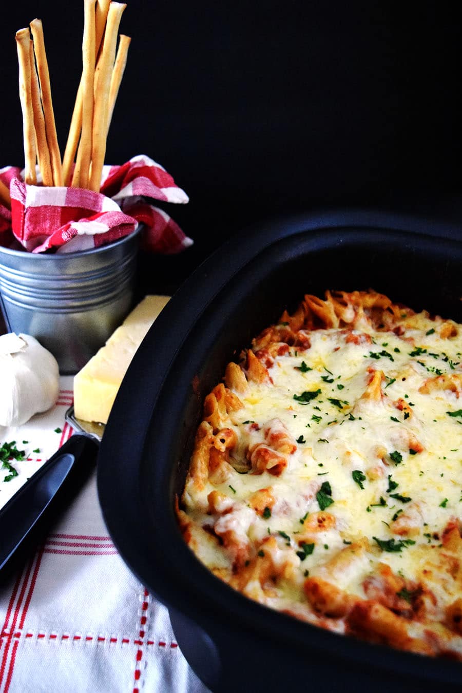 an overhead view of this crock pot chicken parmesan penne pasta with a wedge of parmesan, a cheese grater, and thin bread sticks in the background