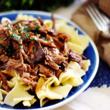 A close up of this crock pot Mississippi pot roast served in a blue bowl over egg noodles