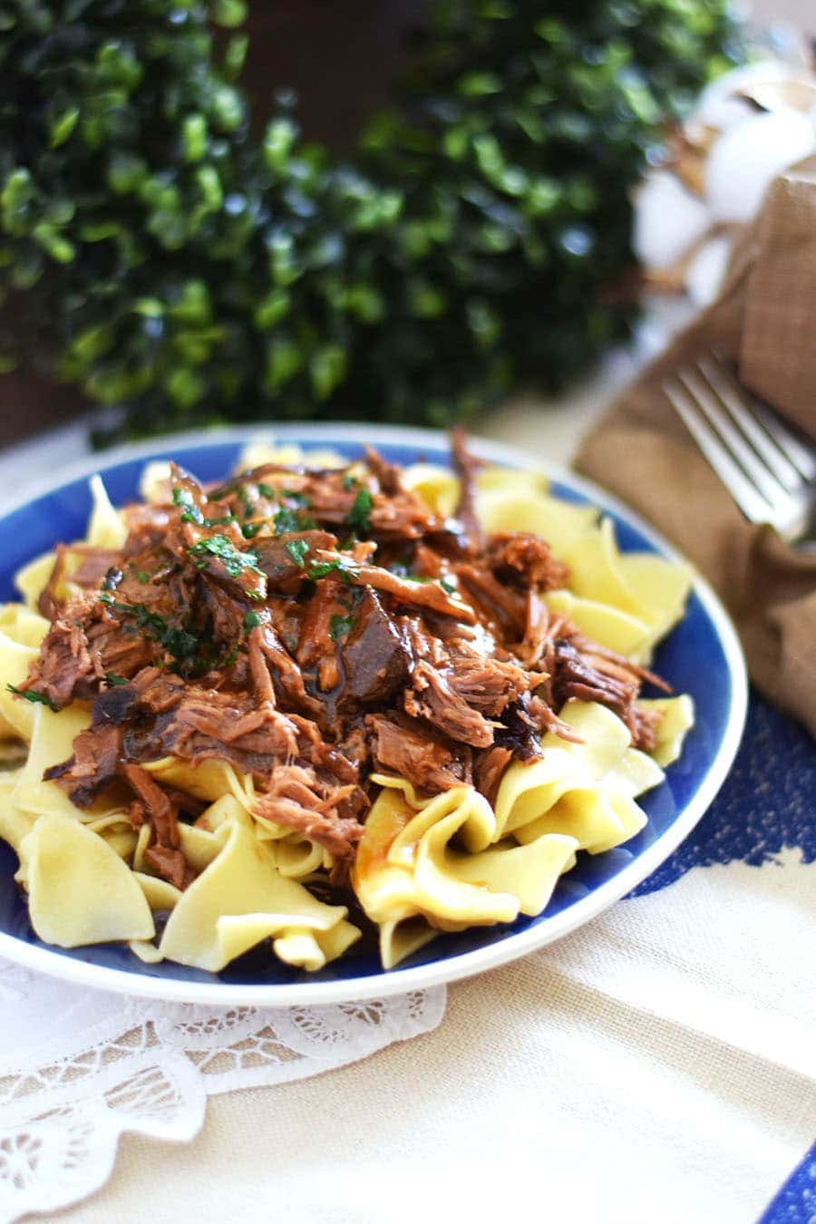 a blue plate with egg noodles and a serving of shredded slow cooker mississippi pot roast
