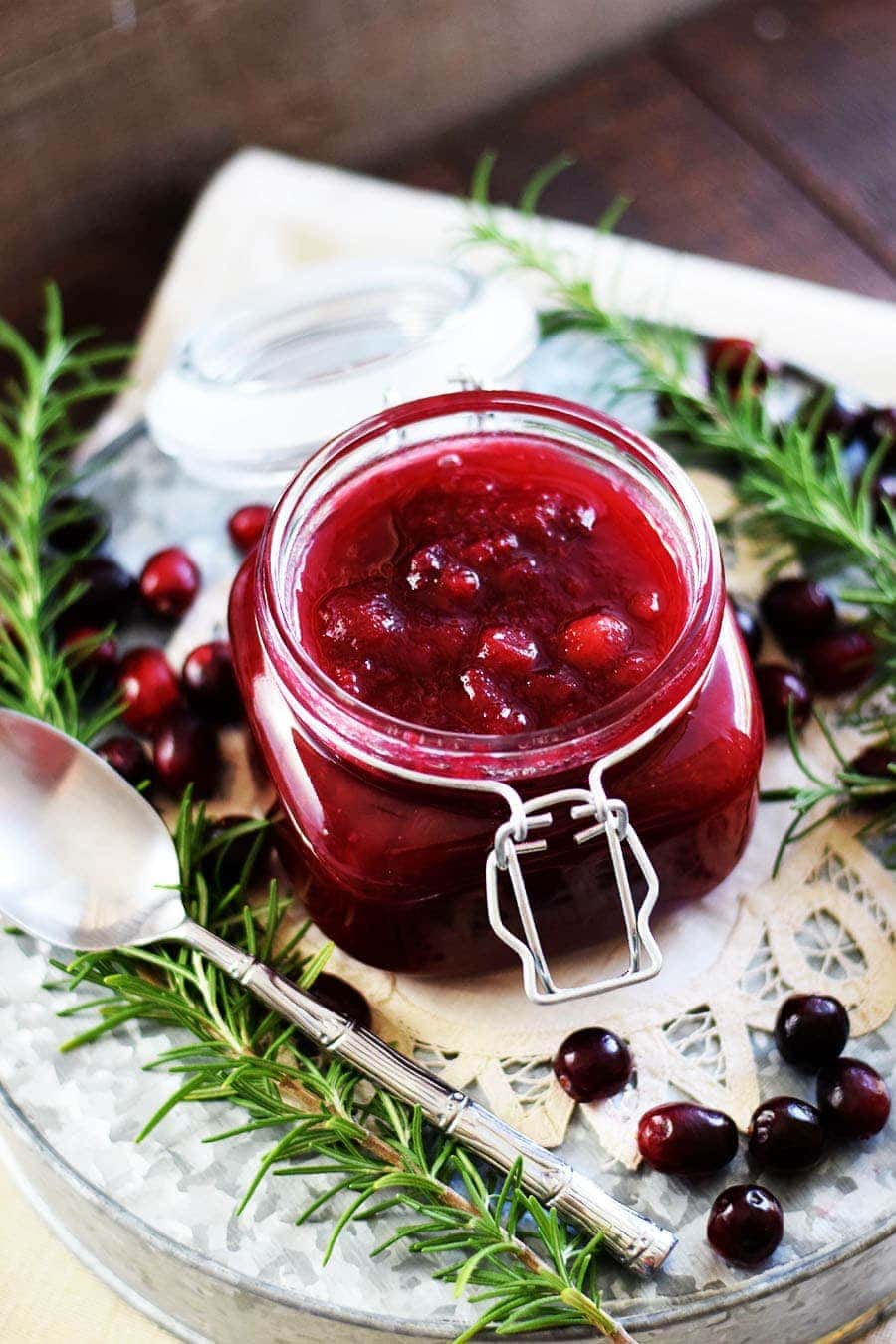 a glass jar  filled with this slow cooker cranberry sauce with rosemary, fresh cranberries, and a silver spoon placed around it