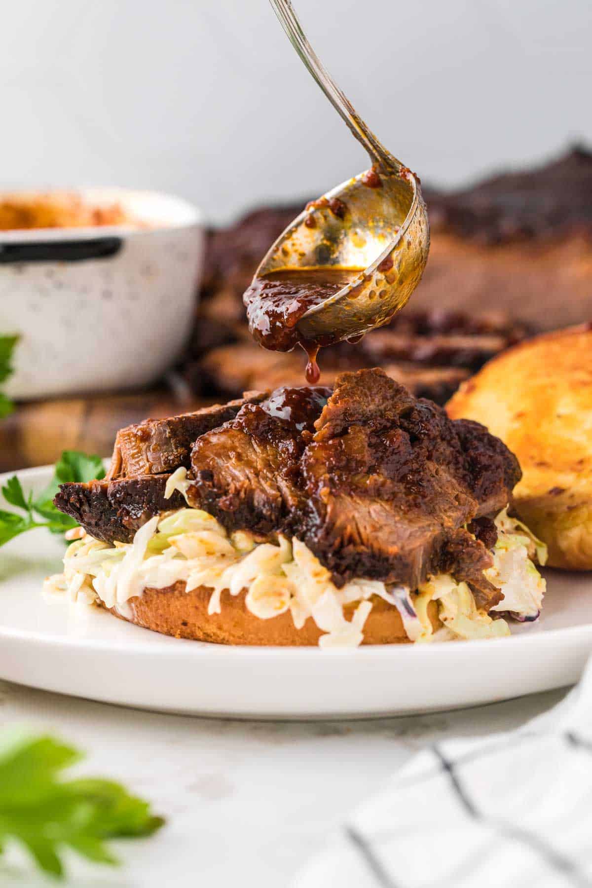 Sliced beef brisket sandwich served with slaw and bbq sauce being drizzled over the top of the meat.