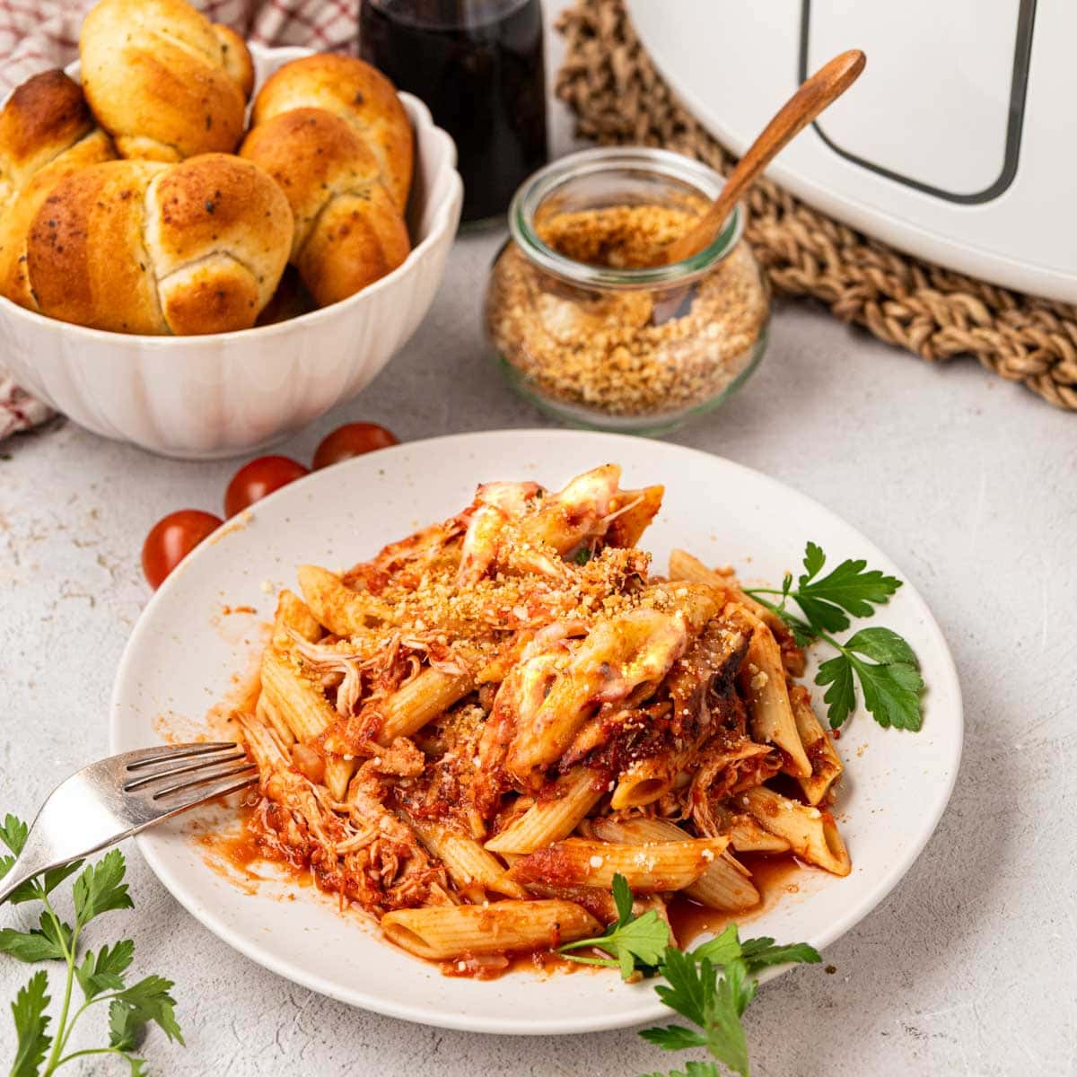 A plate of crockpot chicken parmesan pasta with a garlic bread knot on a white plate.