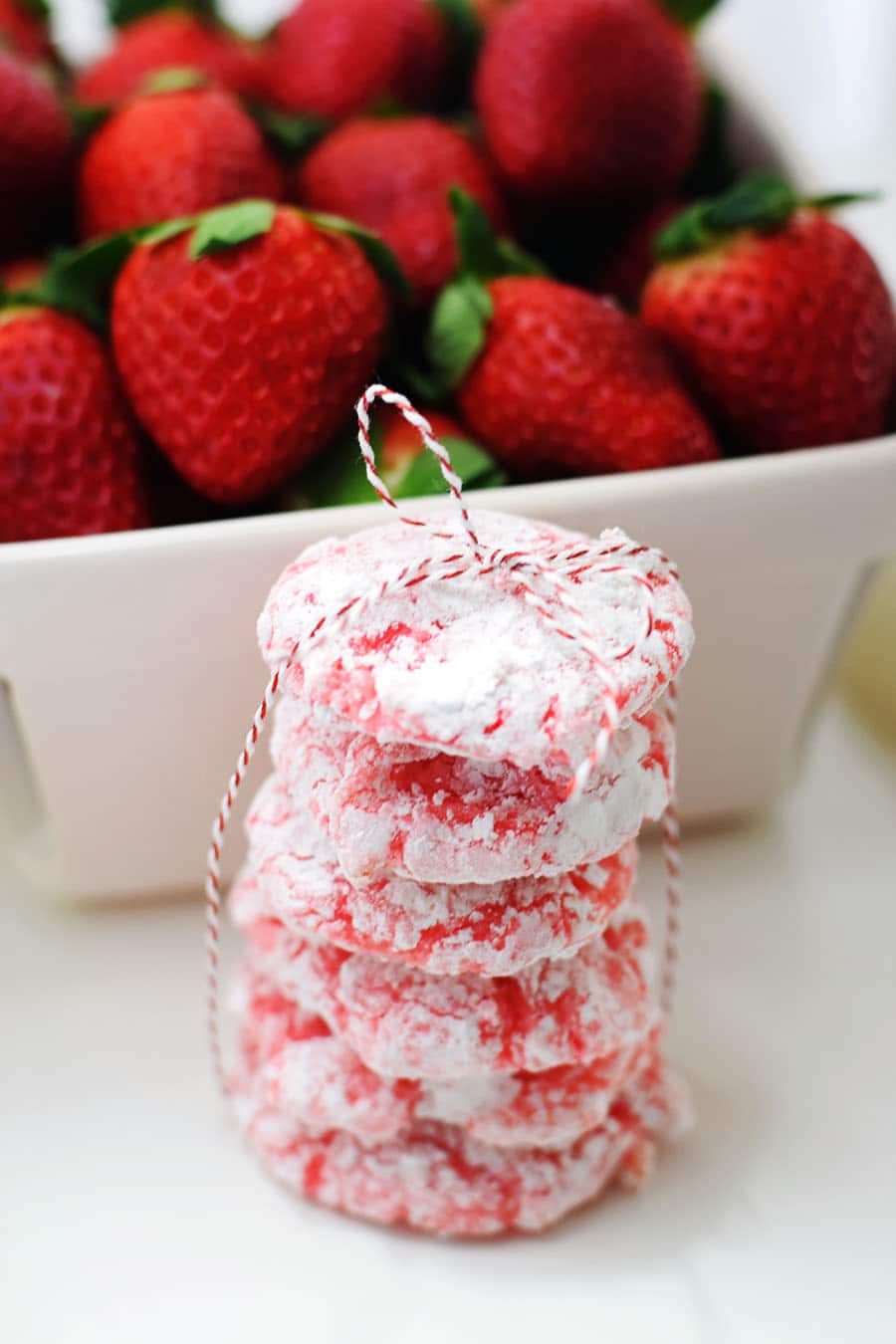 a stack of strawberry crinkle cookies tied together with red and white twine and a basket of fresh strawberries in the background