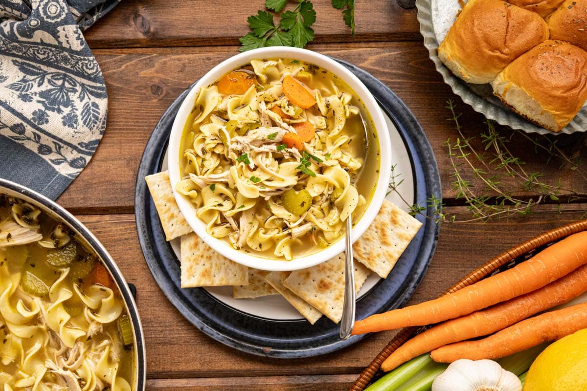 A bowl of chicken noodle soup set on a tablescape with rolls, crackers, and fresh veggies.
