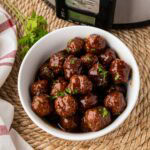 A white bowl filled with strawberry jalapeno pepper jelly meatballs with a crock pot in the background.