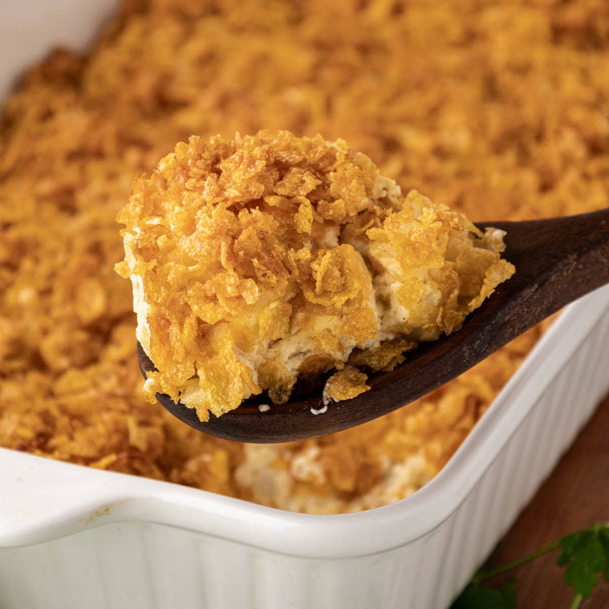 A wooden spoon, removing a scoop of cheesy potato casserole from a baking dish.
