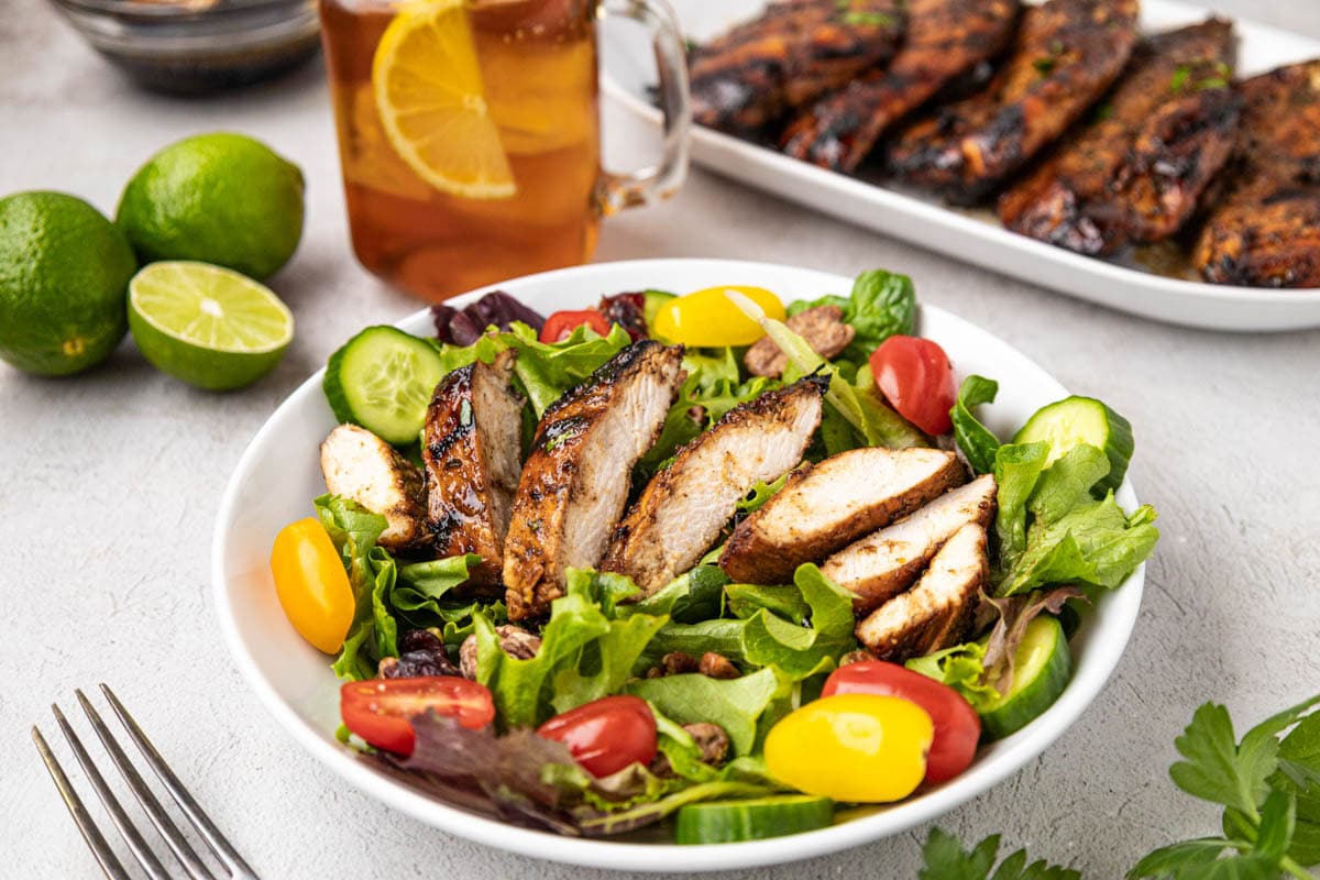 Sliced grilled chicken made in the best grilled chicken marinade over a salad with a platter of the chicken in the background.