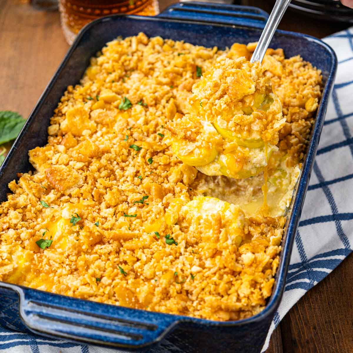 A cheesy yellow squash casserole with a spoon taking a scoop out of the pan.