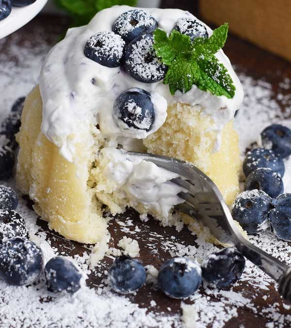 a fork taking a bite out of this almond cake with blueberry whipped cream, powdered sugar, fresh mint, and fresh blueberries