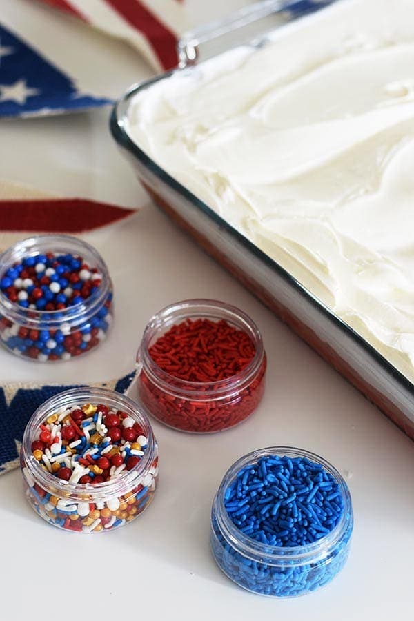 an assortment of red, white, and blue sprinkles alongside this red, white, and blue poke cake for the fourth of july