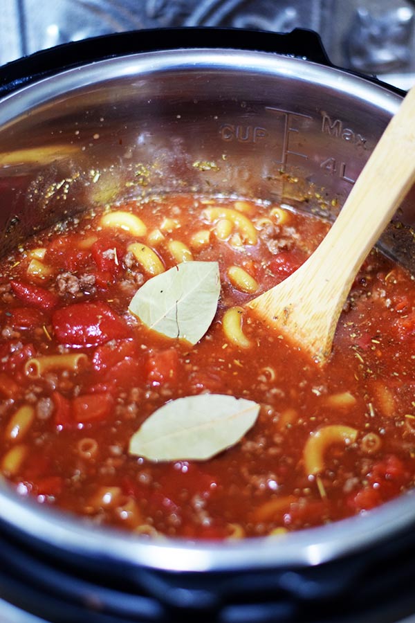 An overhead shot of this instant pot goulash with macaroni noodles, tomatoes, bay  leaves, and a wooden spoon